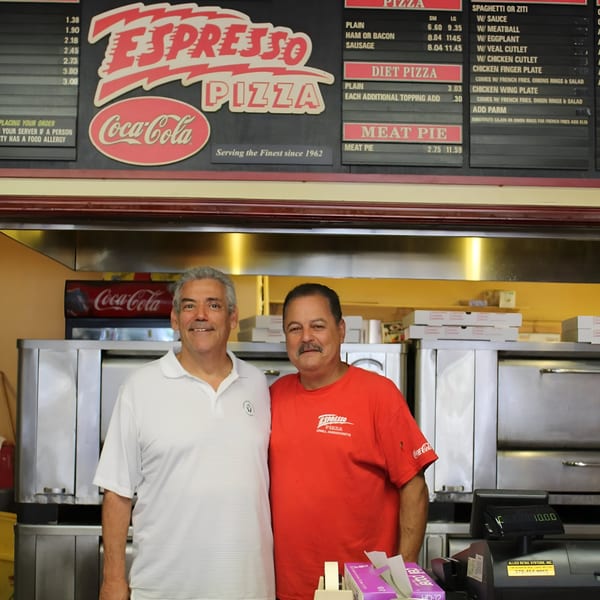 Two men, owners of Espresso Pizza, stand smiling in front of their restaurant's menu board.