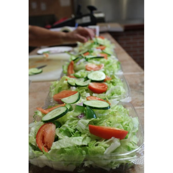 A person prepares multiple fresh garden salads with lettuce, tomatoes, cucumbers, and peppers in plastic containers.