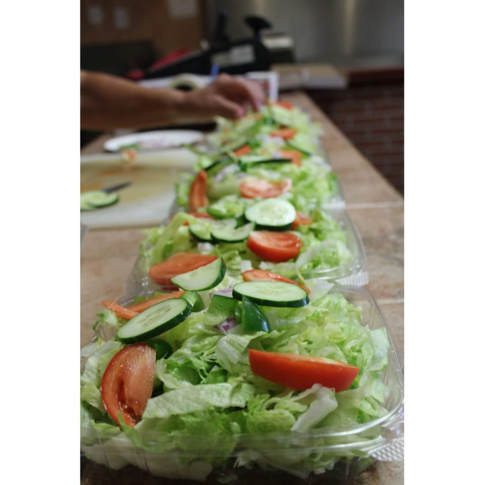 A person prepares multiple fresh garden salads with lettuce, tomatoes, cucumbers, and peppers in plastic containers.