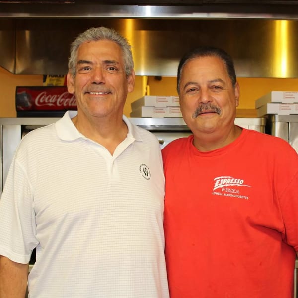 Two smiling men, one in a white polo and one in a red Espresso Pizza shirt, stand in a kitchen.