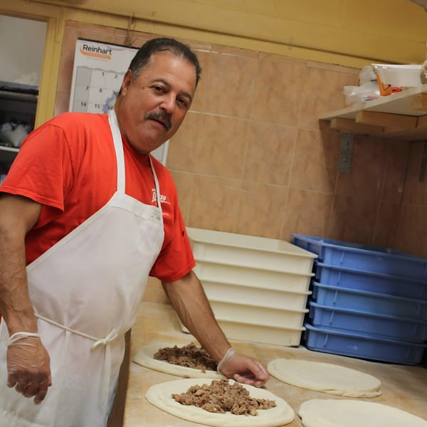 Espresso Pizza chef in a red shirt and white apron preparing pizzas with meat toppings.